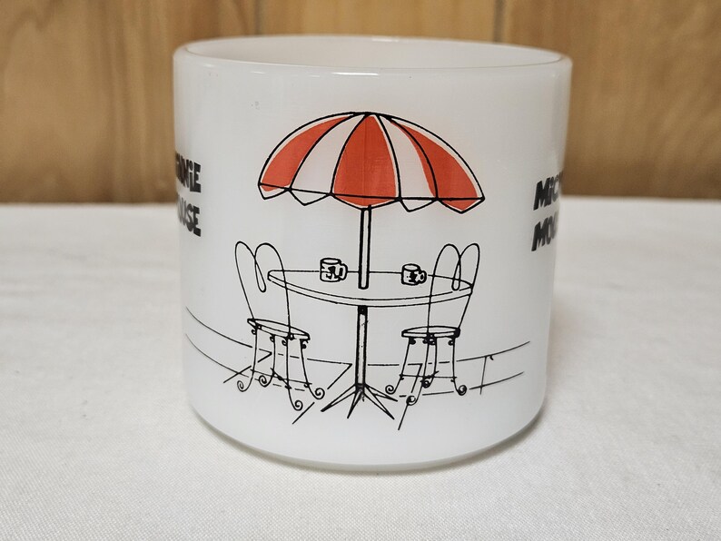 Pu&ograve; includere: Tazza in ceramica bianca con grafica nera e rossa di una scena di caff&egrave;. Il design include un ombrellone a strisce rosse e bianche, un tavolo, due sedie e le parole "Mickey Mouse" sul lato.