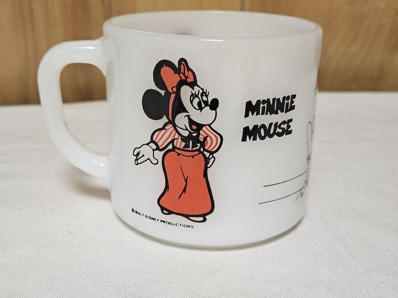 Pu&ograve; includere: Tazza in ceramica bianca con un'illustrazione a cartoni animati di Minnie Mouse con abito e fiocco rossi. Le parole "Minnie Mouse" sono stampate in nero. La tazza ha un manico curvo, probabilmente vintage.