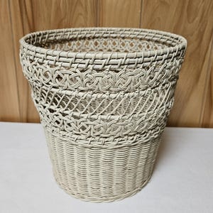 May include: A cream-colored woven basket with a flared shape. The basket features a detailed pattern of interwoven strands, creating a textured surface. The design includes a decorative band around the top and a sturdy base.