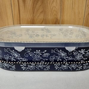 May include: A dark blue oval ceramic baking dish with a white floral vine pattern and two side handles. The dish includes a clear plastic lid with a matching white floral design and a decorative beaded edge along the rim of the base.