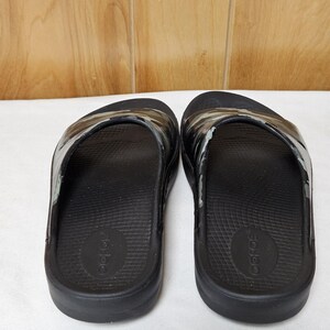 May include: A pair of black slide sandals with a camouflage pattern on the strap. The sandals are made of a rubber-like material and have a contoured footbed. The brand name is visible on the footbed.