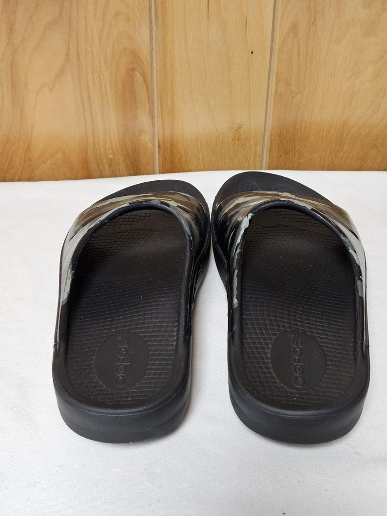 May include: A pair of black slide sandals with a camouflage pattern on the strap. The sandals are made of a rubber-like material and have a contoured footbed. The brand name is visible on the footbed.