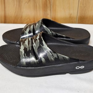 May include: A pair of black slide sandals with a camouflage pattern on the straps. The sandals have a textured footbed and a thick sole. The camouflage design features shades of gray, green, and black. The sandals are designed for comfort and casual wear.