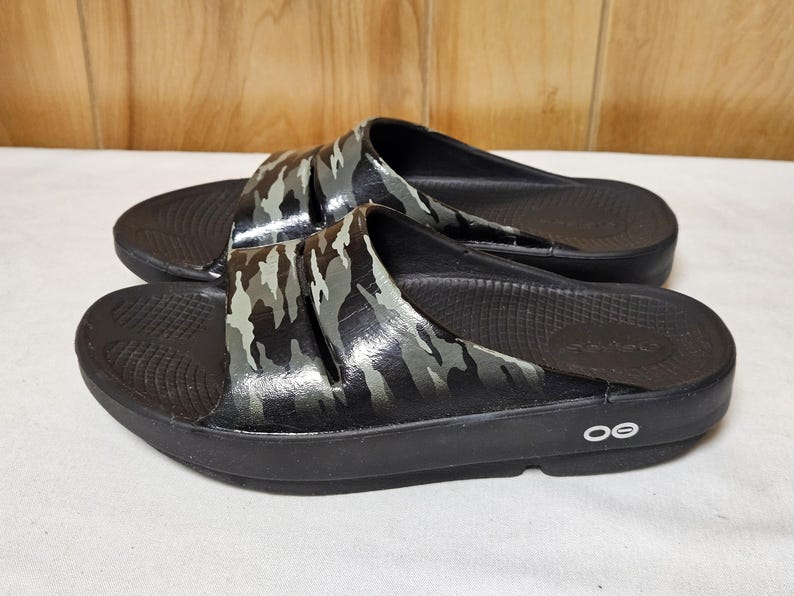 May include: A pair of black slide sandals with a camouflage pattern on the straps. The sandals have a textured footbed and a thick sole. The camouflage design features shades of gray, green, and black. The sandals are designed for comfort and casual wear.