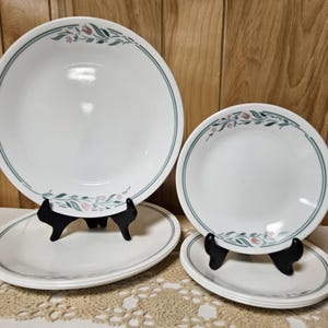 May include: A set of white ceramic dishes with a green border and floral accents. The set includes a large bowl, a smaller plate, and two oval plates. The dishes are displayed on black stands.