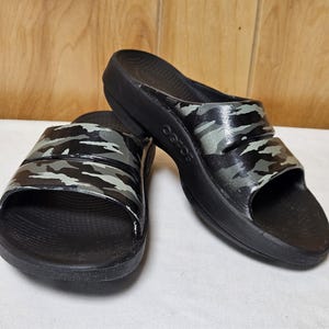 May include: A pair of black slide sandals with a camouflage pattern in shades of gray and green on the straps. The sandals have a thick sole and are displayed on a white surface.