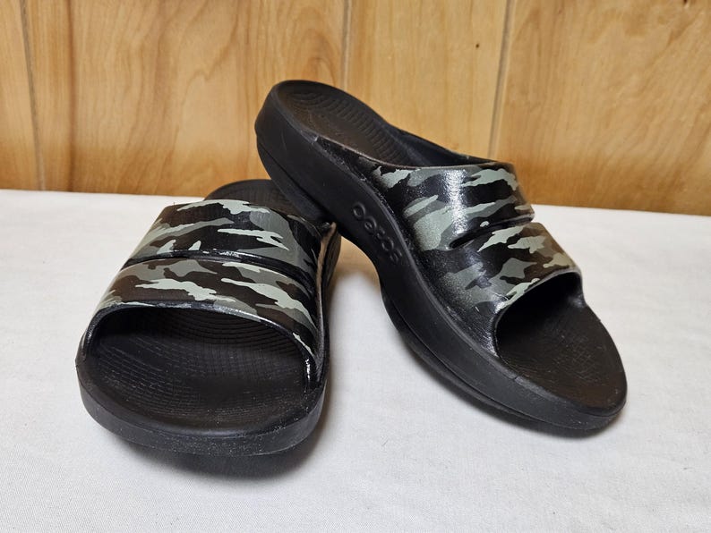 May include: A pair of black slide sandals with a camouflage pattern in shades of gray and green on the straps. The sandals have a thick sole and are displayed on a white surface.