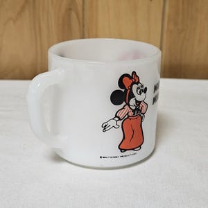 Pu&ograve; includere: Tazza in ceramica bianca con un design a cartoni animati di Minnie Mouse. Minnie &egrave; raffigurata con un vestito e un fiocco rossi, con una camicia a righe. La tazza ha un manico curvo e il testo "Minnie Mouse". Un oggetto da collezione Disney vintage.