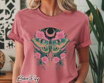 May include: A pink t-shirt with a green and yellow moth design. The moth is surrounded by pink flowers and a crescent moon. The text "BeeJay" is printed on the bottom of the shirt.