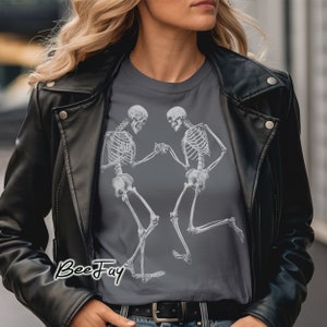 Può includere: Una maglietta grigio scuro con una grafica di due scheletri che ballano. Gli scheletri sono bianchi e si tengono per mano. Il testo "BeeFay" è stampato in nero nella parte inferiore della maglietta.