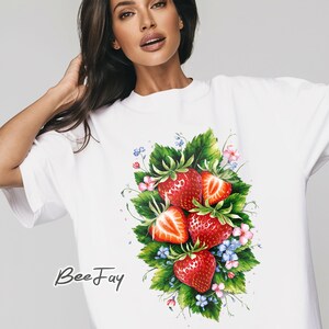 Puede incluir: Camiseta blanca con un diseño de acuarela de fresas maduras, hojas verdes y flores rosas y azules. El texto "Bee Fay" está impreso en la parte inferior izquierda de la camiseta.