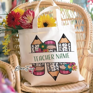 May include: A white canvas tote bag with a pencil and floral design. The design includes pencils with leopard print accents, pink glitter, and colorful flowers. The text "TEACHER NAME" is displayed in green.