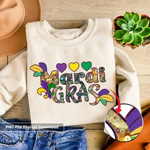 May include: A white sweatshirt featuring the words "Mardi Gras" in purple, green, and gold glitter lettering. The letters are adorned with feathers and beads. The sweatshirt is lying on a wooden surface with a pair of blue jeans and a brown hat.