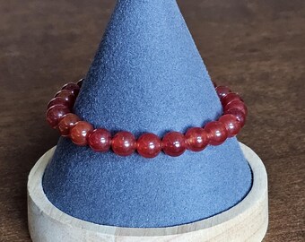 May include: A red beaded bracelet displayed on a blue cone-shaped stand. The bracelet is made of small, round beads. The stand is set on a wooden base, creating a simple yet elegant presentation. The bracelet is a jewelry item.
