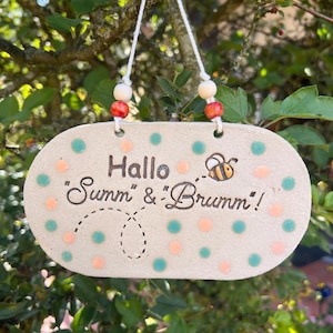Könnte beinhalten: Ein ovales Keramik-Hängeschild in hellem Beige, verziert mit türkisfarbenen und pfirsichfarbenen Punkten. Der Text "Hallo "Summ" & "Brumm"!" ist in Dunkelbraun aufgedruckt, ergänzt durch eine kleine Biene und eine gepunktete Flugbahn.