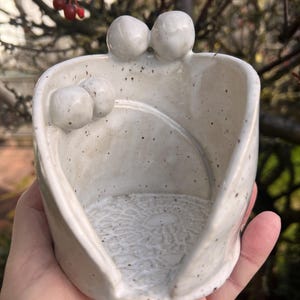 Könnte beinhalten: Ein handgefertigter, cremefarbener Keramik-Löffelhalter mit strukturierter Innenseite und gesprenkelter Oberfläche. Der Löffelhalter hat zwei abgerundete Figuren oben und zwei kleinere Kugeln an der Seite. Das Design ist organisch und künstlerisch.