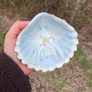 Könnte beinhalten: Eine kleine, handgefertigte Keramikschale mit gewelltem Rand. Die Schale hat eine hellblaue Glasur mit weißen und beigefarbenen Akzenten, die einer Muschel ähnelt. Die Schale wird in der Hand einer Person gehalten, um ihre Größe und Form zu zeigen.