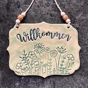 Könnte beinhalten: Ein Keramik-Willkommensschild mit dem Wort "Willkommen" in türkisfarbener Schrift. Das Schild zeigt ein Blumendesign in Grüntönen und wird an einer weißen Schnur mit Holzperlen aufgehängt. Das Schild hat eine Craquelé-Glasur.