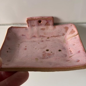 Könnte beinhalten: Eine handgefertigte Keramik-Seifenschale mit rechteckiger Form und Griff. Die Schale ist in Rosa- und Brauntönen glasiert und hat eine gesprenkelte Textur. Die Ränder sind dunkler braun.