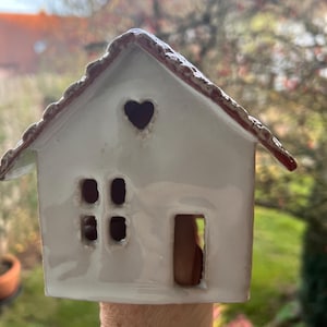 Könnte beinhalten: Ein weißes Keramikhaus mit einem herzförmigen Fenster und vier quadratischen Fenstern. Das Dach ist mit einem rotbraunen Rand versehen. Das Haus hat eine rechteckige Tür und wird von einer Person gehalten.