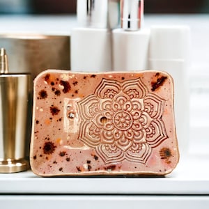 Könnte beinhalten: Eine rosa Keramik-Seifenschale mit einem goldenen Mandala-Design. Die Schale hat eine gesprenkelte Glasur und einen leicht erhöhten Rand.