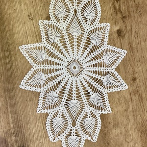 Könnte beinhalten: Weißer, handgefertigter Häkel-Tischläufer mit detailliertem Lochmuster. Der Läufer hat ein zentrales kreisförmiges Motiv mit strahlenförmigen Mustern und spitzen, blattartigen Kanten. Die filigrane, spitzenartige Textur wird auf einer Holzoberfläche dargestellt.
