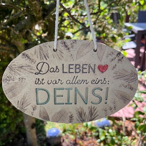 Könnte beinhalten: Ovale Keramikschild mit dem deutschen Text "Das Leben ist vor allem eins: DEINS!" in schwarzer und türkisfarbener Schrift. Das Schild hat einen beigen Hintergrund mit Blättermustern und einem roten Herz. Es hängt an einer weißen Schnur.