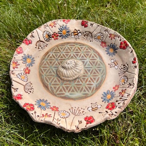 Könnte beinhalten: Handgefertigtes Vogelbad aus Keramik mit floralem Design. Die beige Schale zeigt rote, blaue und weiße Blumen sowie Bienen. Die Mitte hat ein geometrisches Muster und ein erhabenes, strukturiertes Mittelstück.