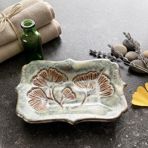 Könnte beinhalten: Eine Keramik-Seifenschale mit einem Ginkgo-Blatt-Design in Braun und Weiß. Die Schale ist rechteckig mit einem gewellten Rand. Eine grüne Flasche, Handtücher und ein gelbes Blatt sind ebenfalls sichtbar.