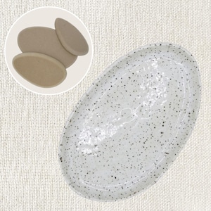 Könnte beinhalten: Ovale, weiße Keramik-Servierplatte mit schwarzen Sprenkeln und leicht erhöhtem Rand. Ein kleineres Bild zeigt drei ovale, beige Untersetzer.