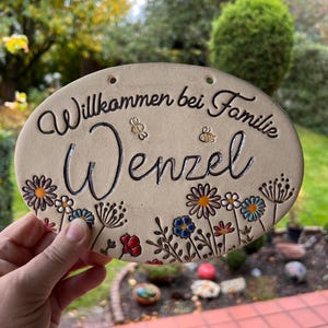 Könnte beinhalten: Ein ovales Keramikschild mit cremefarbener Oberfläche. Der Text "Willkommen bei Familie Wenzel" ist in Schwarz eingraviert. Das Design zeigt handbemalte, bunte Blumen in Rot-, Blau- und Orangetönen sowie zwei kleine Bienen.
