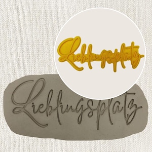 Könnte beinhalten: Ein gelber Plastik-Ausstecher mit dem Text "Lieblingsplatz" in einer kursivschriftlichen Schriftart. Der Ausstecher wird auf einer grauen Oberfläche gezeigt, in die der Text eingeprägt ist.