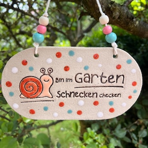 Könnte beinhalten: Ein ovales Keramikschild in hellem Beige, verziert mit kleinen roten, weißen und blauen Punkten. Es zeigt eine Schnecken-Illustration und den Text "Bin im Garten Schnecken checken". Das Schild hängt an einer weißen Schnur mit bunten Holzperlen.