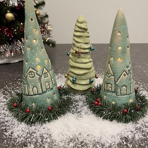 Könnte beinhalten: Drei keramische Weihnachtsbaumdekorationen. Zwei sind türkis, kegelförmig mit Hausdesigns und Lichtern. Der dritte ist ein kleinerer, grüner, spiralförmiger Baum mit Ornamenten.