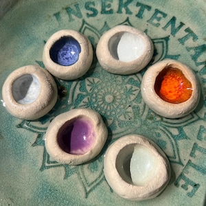 Könnte beinhalten: Eine Sammlung von sieben kleinen, handgefertigten Keramikstücken, jedes mit einem einzigartigen farbigen Innenraum: blau, weiß, orange, lila und weiß. Die Stücke sind auf einer türkisfarbenen Keramikplatte mit einem floralen Design und dem Wort "INSEKTENT" in Grün angeordnet.