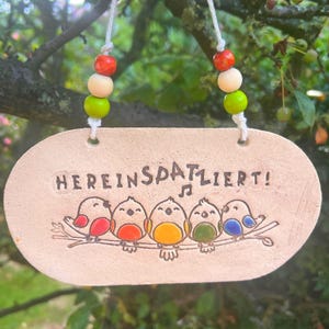 Könnte beinhalten: Ein ovales Keramikschild mit der Aufschrift "HEREINSPAZIERT!" und einer Grafik von fünf bunten Vögeln auf einem Ast. Das Schild hängt an einer weißen Schnur, die mit roten, beigen und lindgrünen Holzperlen verziert ist und ein Notensymbol zeigt.