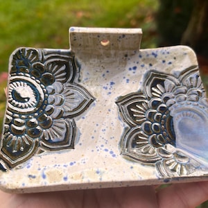 Könnte beinhalten: Eine rechteckige Seifenschale aus Keramik mit Griff. Die Schale ist cremefarben mit blauen Sprenkeln und weist zwei geprägte Blumendesigns in Blau- und Brauntönen auf. Die Schale wird in einer Hand gehalten.