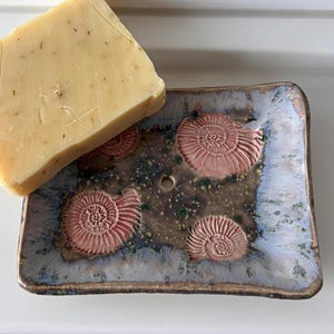 Könnte beinhalten: Rechteckige Keramik-Seifenschale mit blau, braun und grün glasierter Oberfläche, verziert mit vier erhabenen rosa Ammoniten-Fossilien-Designs. Eine hellfarbene Seife liegt auf der Schale. Ein dekoratives Badezimmer-Accessoire.