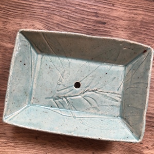 Könnte beinhalten: Eine hellblaue Keramik-Seifenschale mit einem Blattdesign und einem Abflussloch.