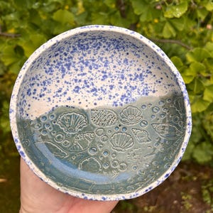 Könnte beinhalten: Eine runde Keramikschale mit zweifarbigem Design. Die obere Hälfte ist weiß mit blauen Sprenkeln, während die untere Hälfte eine einfarbige blaugraue Glasur aufweist, die mit verschiedenen Muschelmustern, einschließlich Jakobsmuscheln, geprägt ist.