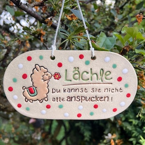 Könnte beinhalten: Ovales Keramikschild mit einer Lama-Illustration, dem deutschen Wort "Lächle" und dem Satz "Du kannst sie nicht alle anspucken!" in grüner Schrift. Das Schild hat rote, grüne und weiße Punkte und wird von einer weißen Schnur gehalten.