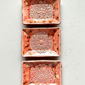 Könnte beinhalten: Drei rechteckige Seifenschalen aus Keramik, vertikal angeordnet. Jede Schale ist korallenfarben mit einem einzigartigen geprägten Blumen- oder geometrischen Design. Die Schalen haben ein gesprenkeltes, rustikales Aussehen.
