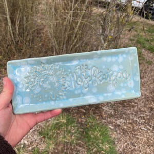 Könnte beinhalten: Ein rechteckiges, hellblaues Keramiktablett mit einem Mandala-Design und dem Text "Für Naschkatzen". Das Tablett wird in einer Hand gehalten und hat eine glänzende Oberfläche. Das Tablett ist etwa 25 cm lang.