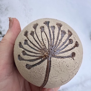 Könnte beinhalten: Runder, beigefarbener Keramikgegenstand mit einem dunkelbraunen Blumendesign. Das Design ähnelt einem Löwenzahn oder einer ähnlichen Pflanze, mit einem zentralen Stiel und radialen Linien. Der Gegenstand wird in einer Hand vor einem schneebedeckten Hintergrund gehalten.