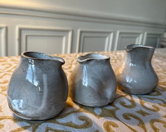 May include: Three small, white ceramic pitchers with a gray glaze. The pitchers have a rounded shape and a small spout. They are arranged in a row on a white and gold patterned tablecloth.