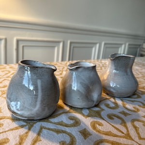 May include: Three small, white ceramic pitchers with a gray glaze. The pitchers have a rounded shape and a small spout. They are arranged in a row on a white and gold patterned tablecloth.