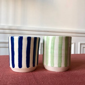 May include: Two ceramic mugs with white and blue stripes and white and green stripes. The mugs are sitting on a red surface.