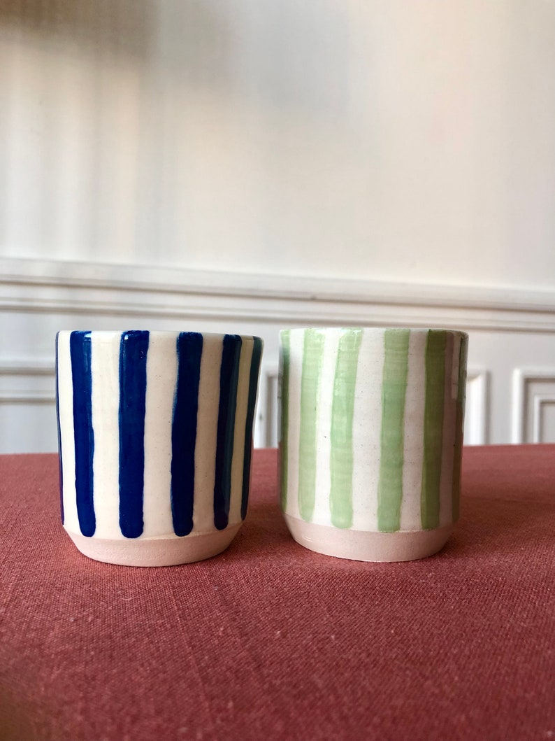 May include: Two ceramic mugs with white and blue stripes and white and green stripes. The mugs are sitting on a red surface.