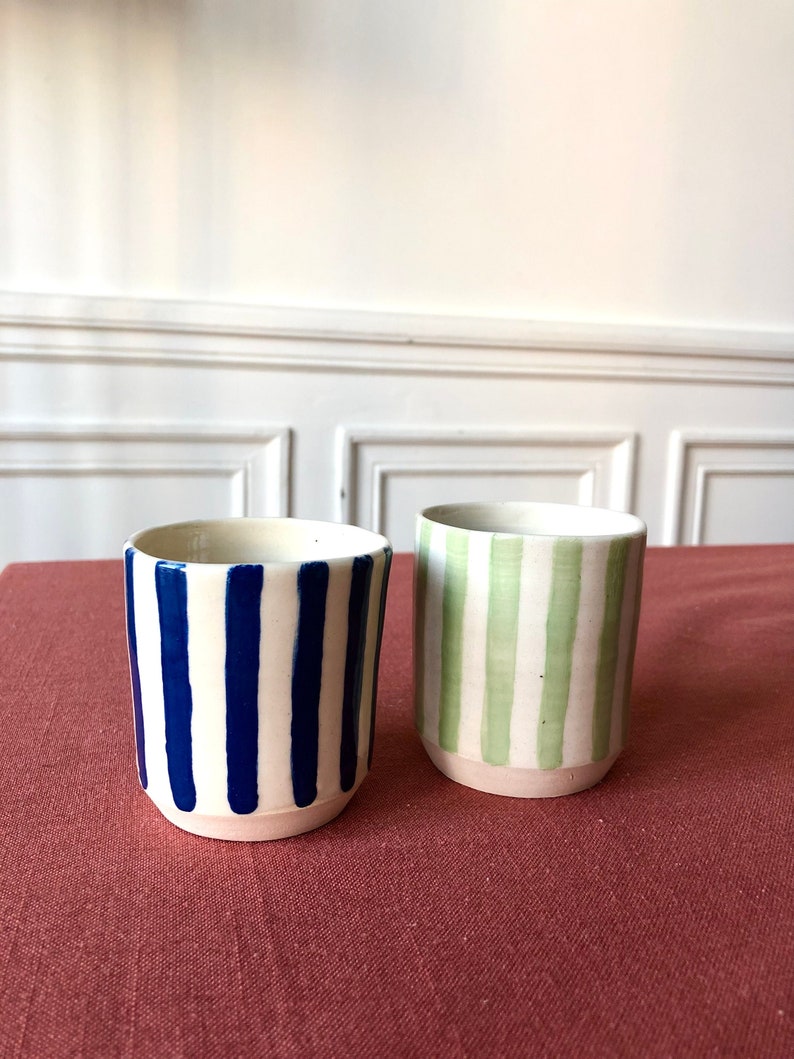 May include: Two ceramic mugs with striped patterns. One mug has blue stripes on a white background, and the other has green stripes on a white background.