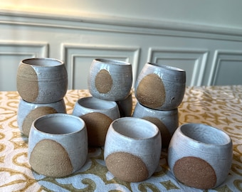 May include: A set of ten small ceramic cups with a white glaze and a brown speckled design. The cups are stacked in a pyramid shape on a white and gold patterned tablecloth.